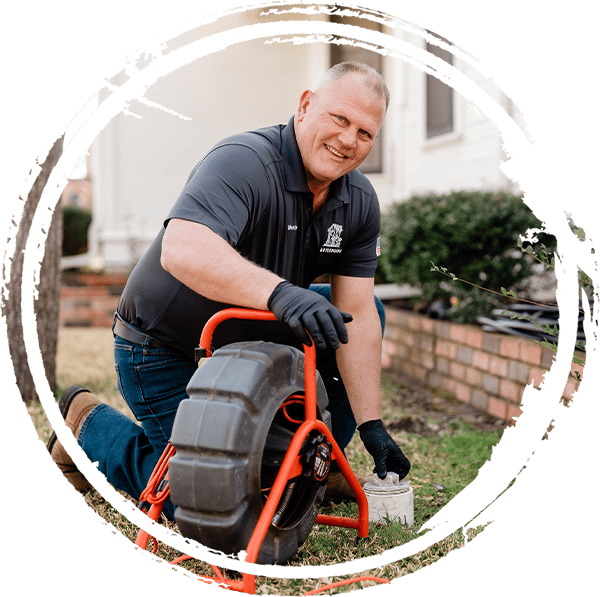 Round photo of S & D plumber smiling while working on a pipe drain outside a home.